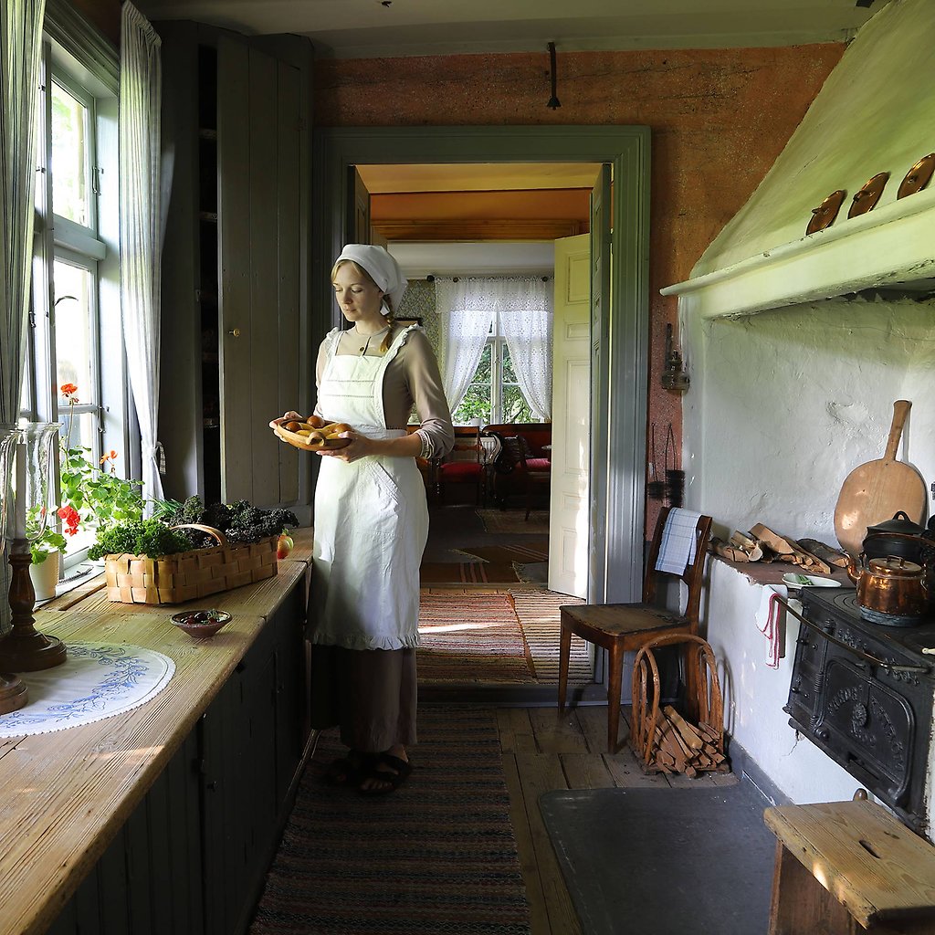 En kvinna i historiska kläder står i ett gammaldags kök